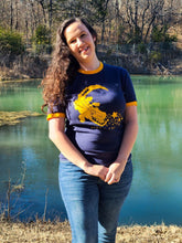 Model is wearing a size Large Croc Star ringer t-shirt.  It is a blue shirt with yellow rings on the collar and sleeve openings. She is posing with her hands in front standing by a lovely lake.