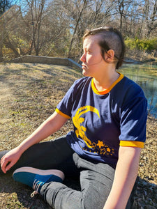 Model is wearing an XL Croc Star ringer t-shirt. It is a blue shirt with yellow rings on the collar and sleeve openings. She is sitting on the ground next to a lovely lake.