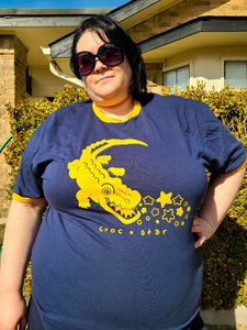 Model is wearing a 2XL Croc Star ringer t-shirt. It is a blue shirt with yellow rings on the collar and sleeve openings. They are posing with their arm on their hip wearing cool sunglasses.